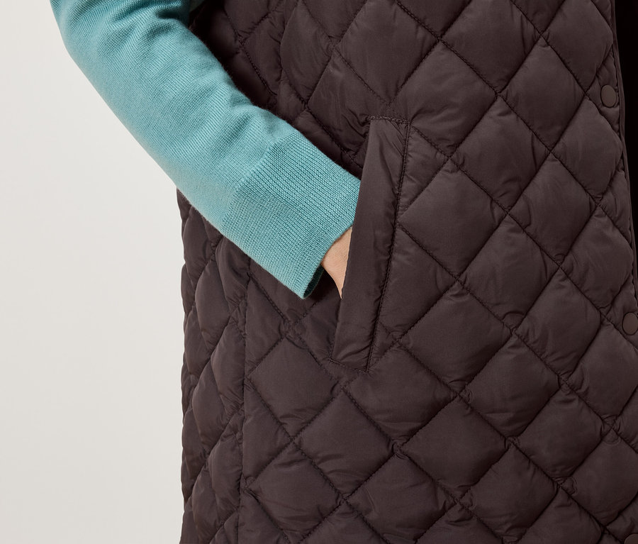 Hand in der Tasche einer gesteppten, braunen Weste, unter der Weste ist ein blauer Pullover zu sehen.