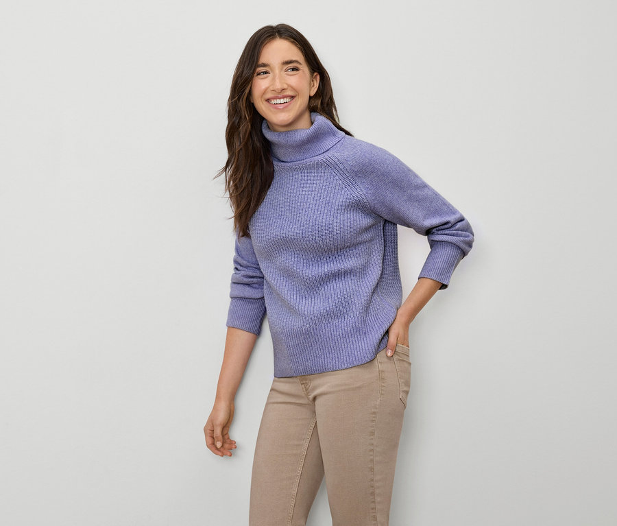 Lächelnde Frau posiert in lila Rollkragenpullover und beigen Hosen.