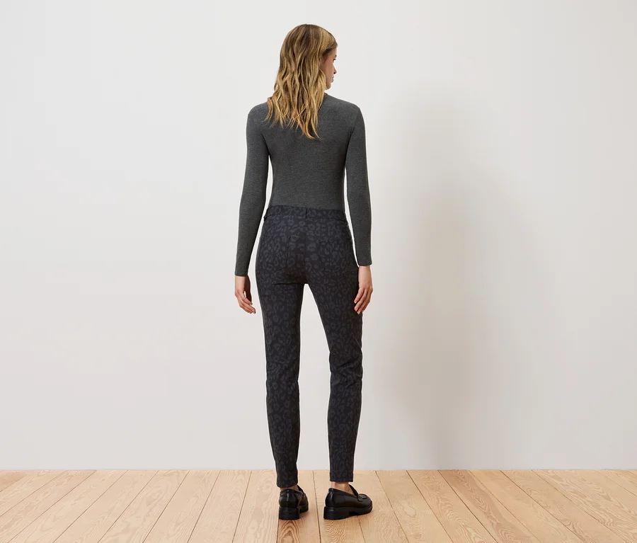 Frau steht mit dem Rücken zur Kamera und trägt ein graues Langarmshirt und eine schwarze gemusterte Hose.