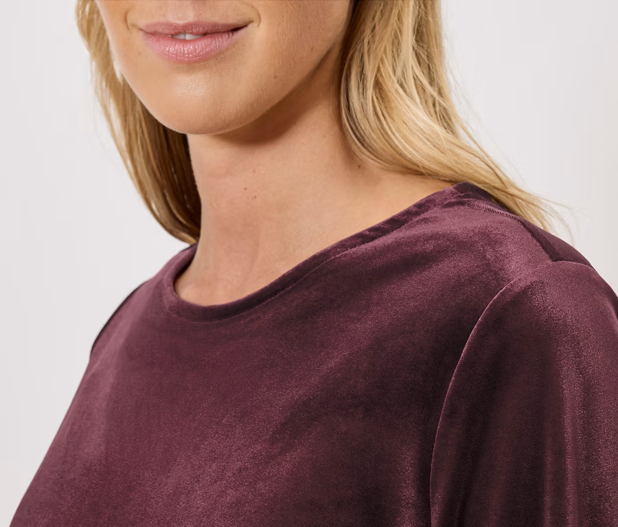 Detailaufnahme einer blonden Frau, die ein weinrotes Samtlangarmshirt trägt.