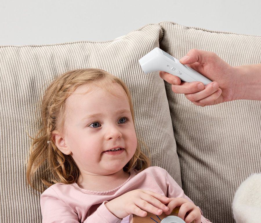 Kleines Mädchen liegt auf einem Sofa, während eine Hand ein weißes, digitales Thermometer an ihre Stirn hält.