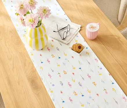 Auf einem Holztisch liegt ein Tischläufer mit Blumenmuster, darauf eine gelb-weiß gestreifte Vase mit rosa Blumen, eine Zeitung mit Brille, Kekse und eine rosa Tasse.