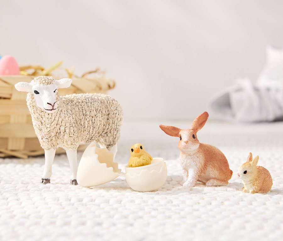 Ein Schaf steht neben einer zerbrochenen Eierschale, aus der ein Küken lugt, daneben sitzen ein Hase und ein kleiner Hase. Im Hintergrund befindet sich ein Weidenkorb mit Süßigkeiten und weiße Turnschuhe. Schleich® Set »Ostertiere«.