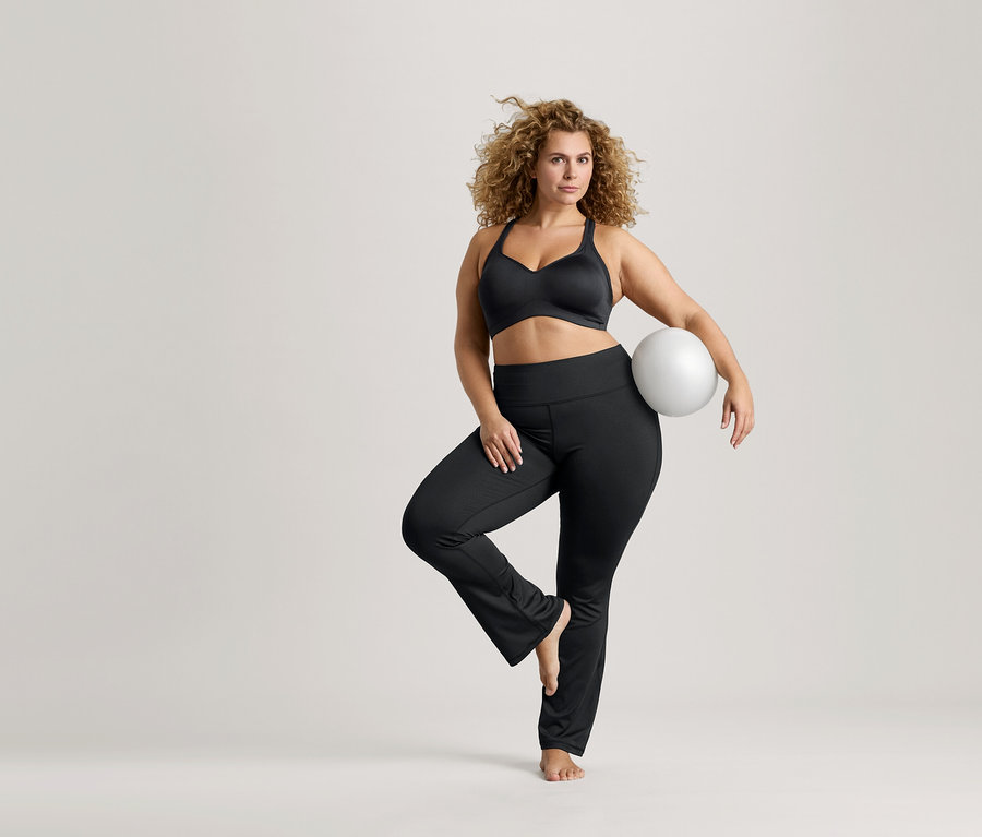 Frau mit lockigem Haar balanciert auf einem Bein, hält einen weißen Ball, trägt einen schwarzen Sport-BH und schwarze Leggings.