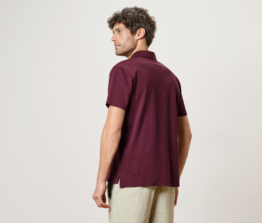 Ein Mann mit lockigem Haar, von hinten gesehen, trägt ein burgunderfarbenes Poloshirt und beigefarbene Shorts.