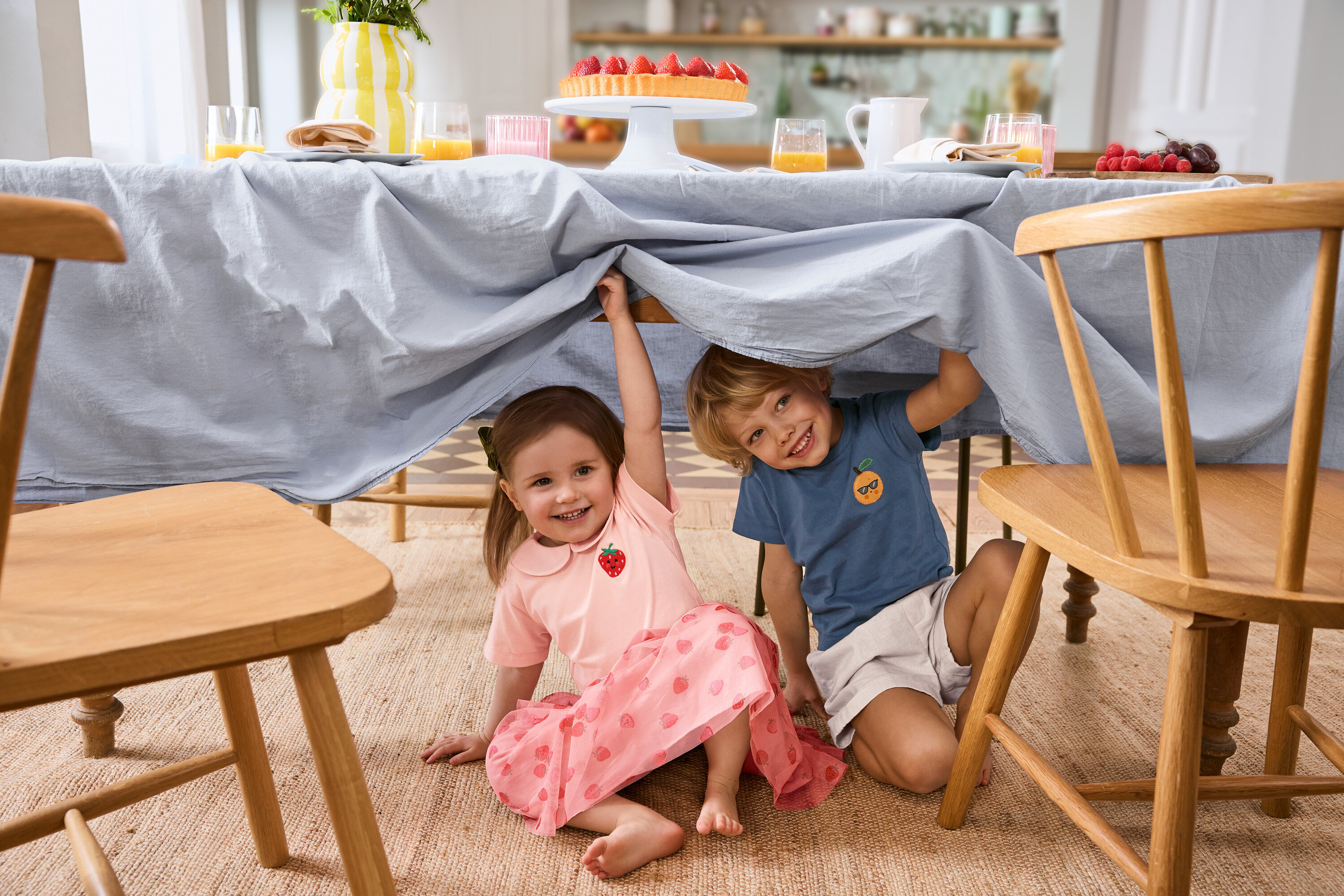Zwei Kinder sitzen unter einem Tisch mit hellblauer Decke. Das Mädchen trägt einen Kinder-Tüllrock und ein 2 Kinder-T-Shirts. Der Junge trägt Kinder-Shorts.
