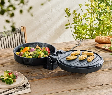 Gemüse in einer schwarzen Pfanne und Fleisch auf einem Grill auf einem Holztisch. Teller mit Gemüse, Brot und Kräuterbutter im Hintergrund.