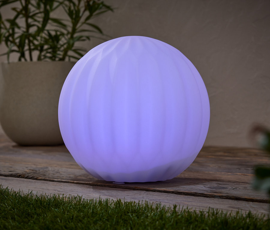 Eine runde, lila Lampe steht auf einem Holztisch, neben einer Topfpflanze.