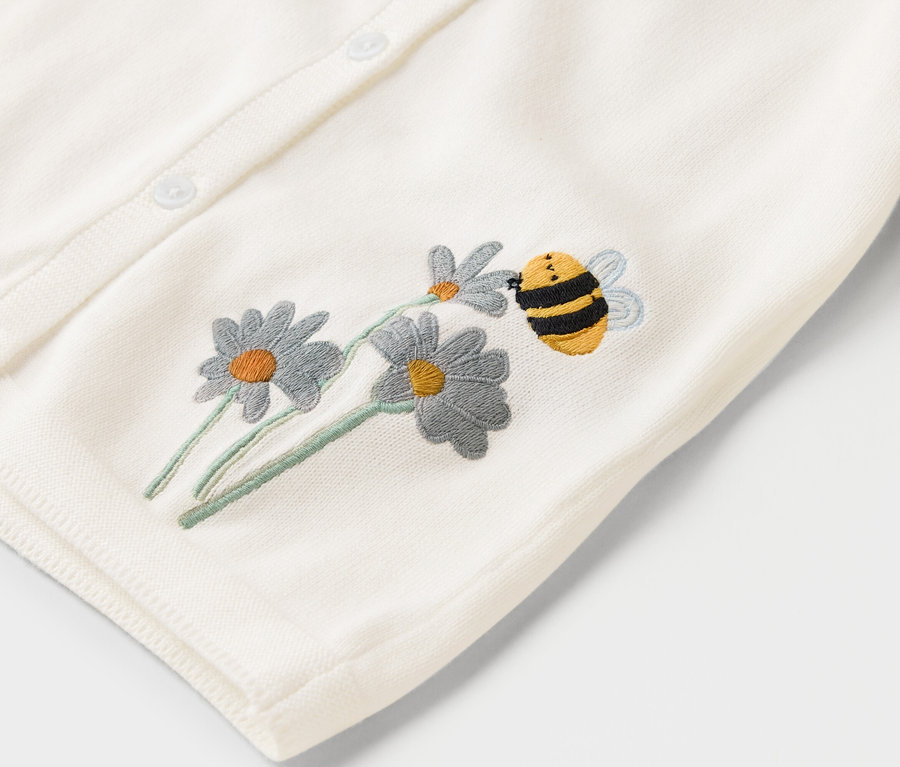 Nahaufnahme einer cremefarbenen Strickjacke mit Knöpfen, bestickt mit Gänseblümchen und einer summenden Biene.