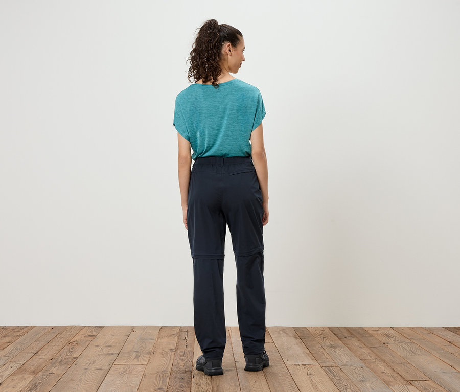 Frau steht mit dem Rücken zur Kamera, trägt ein blaues T-Shirt und eine dunkle Hose mit abzippbaren Hosenbeinen.