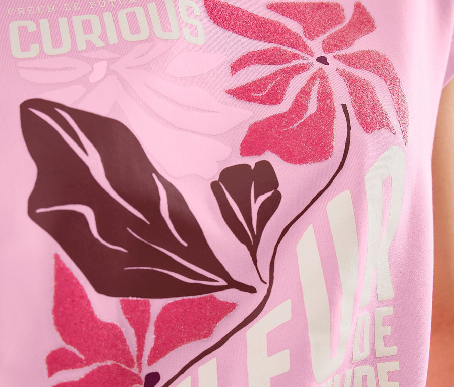 Eine Person trägt ein rosa CECIL Placement-Shirt mit Blumenmuster in Pink und Dunkelbraun und den Schriftzügen "CURIOUS" und "FLEUR DE VIVRE".