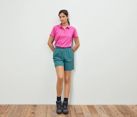 Eine Frau steht vor einer weißen Wand und trägt ein rosa Poloshirt und grüne Shorts.