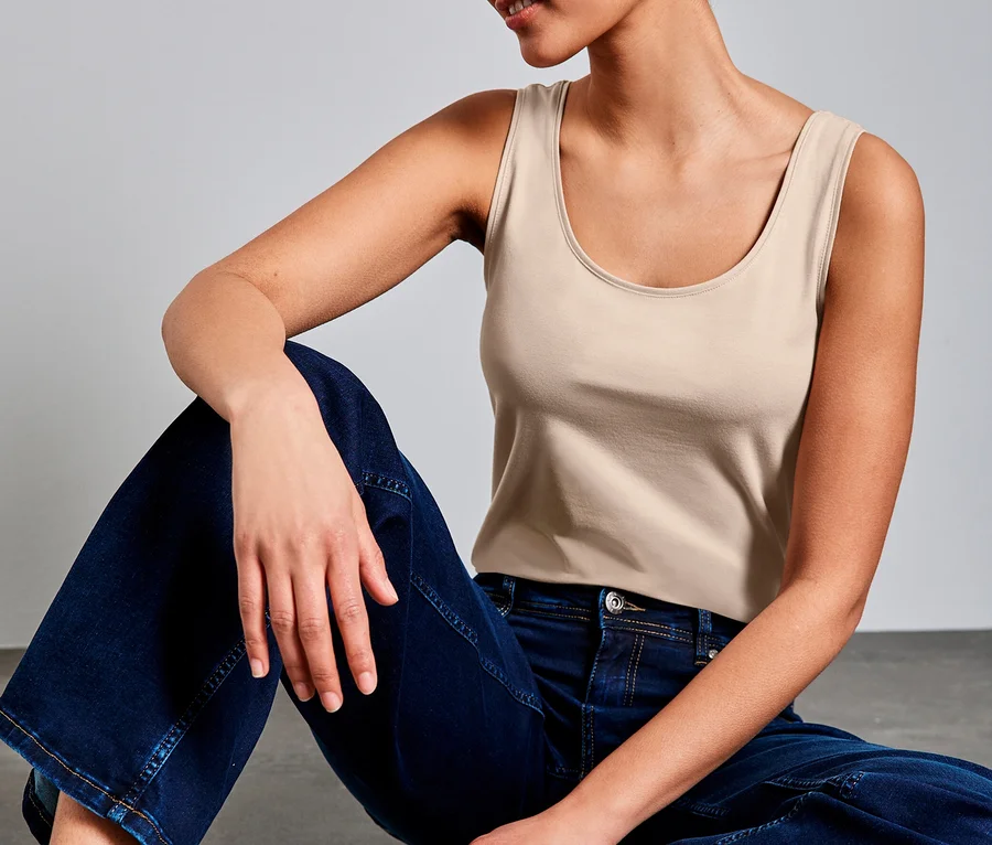 Frau mit kurzen Locken sitzt auf grauem Boden und trägt ein beiges Street One Basic-Top und weite Jeans.