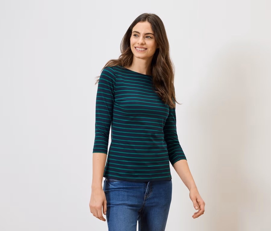 Frau mit langen braunen Haaren trägt ein grün-schwarz gestreiftes Langarmshirt und Jeans vor einem weißen Hintergrund.