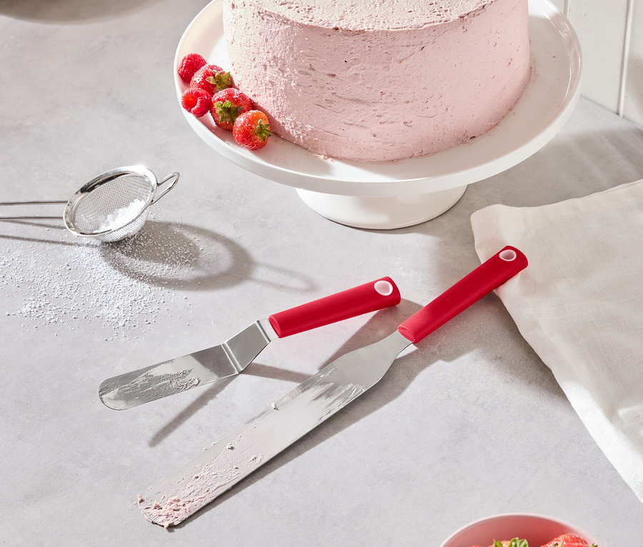 Ein rosa Kuchen mit Erdbeeren und Himbeeren steht auf einem weißen Tortenständer, zusammen mit zwei roten Spateln, einer Schale Erdbeeren und einem Puderzuckersieb.