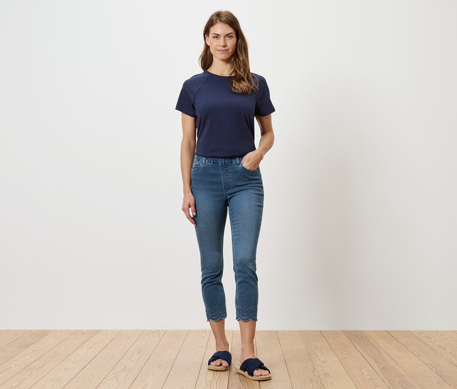 Frau in marineblauem T-Shirt und blauen Jeans mit Lochstickerei am Saum, posiert vor einer weißen Wand.