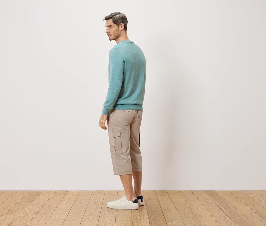 Ein Mann steht seitlich und trägt einen türkisfarbenen Pullover, beigefarbene Shorts und weiße Schuhe.