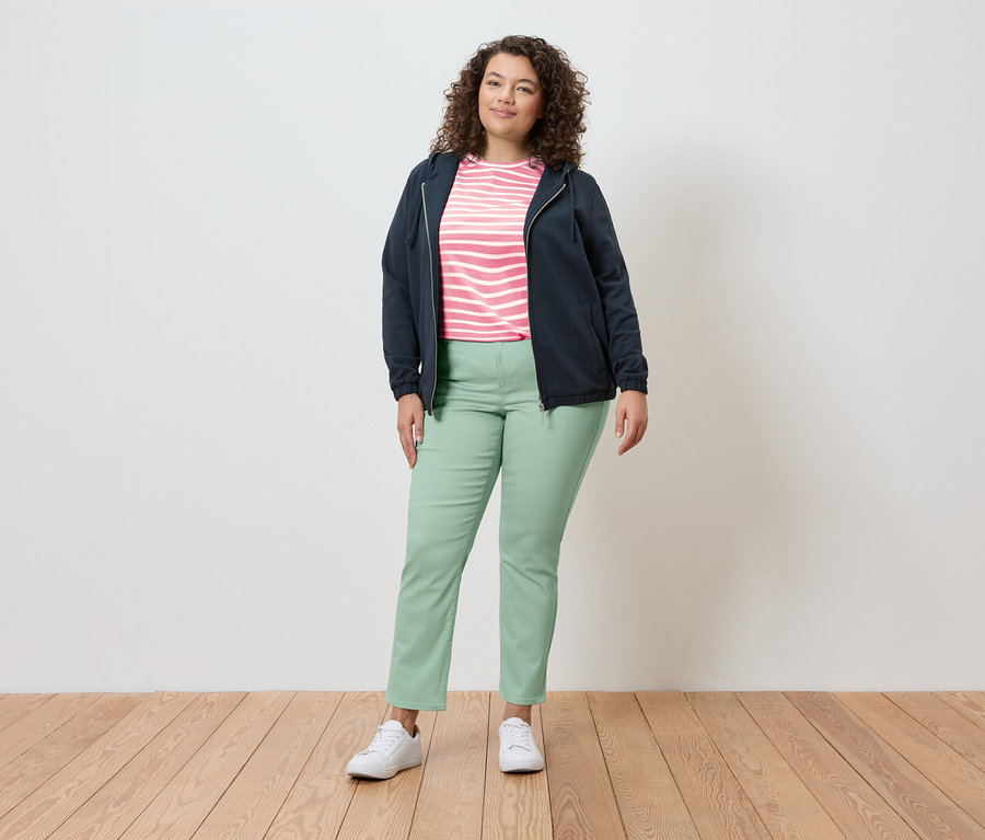 Eine Frau steht in einer hellen Umgebung und trägt eine marineblaue Jacke, ein gestreiftes Oberteil, hellgrüne Hosen und weiße Turnschuhe.