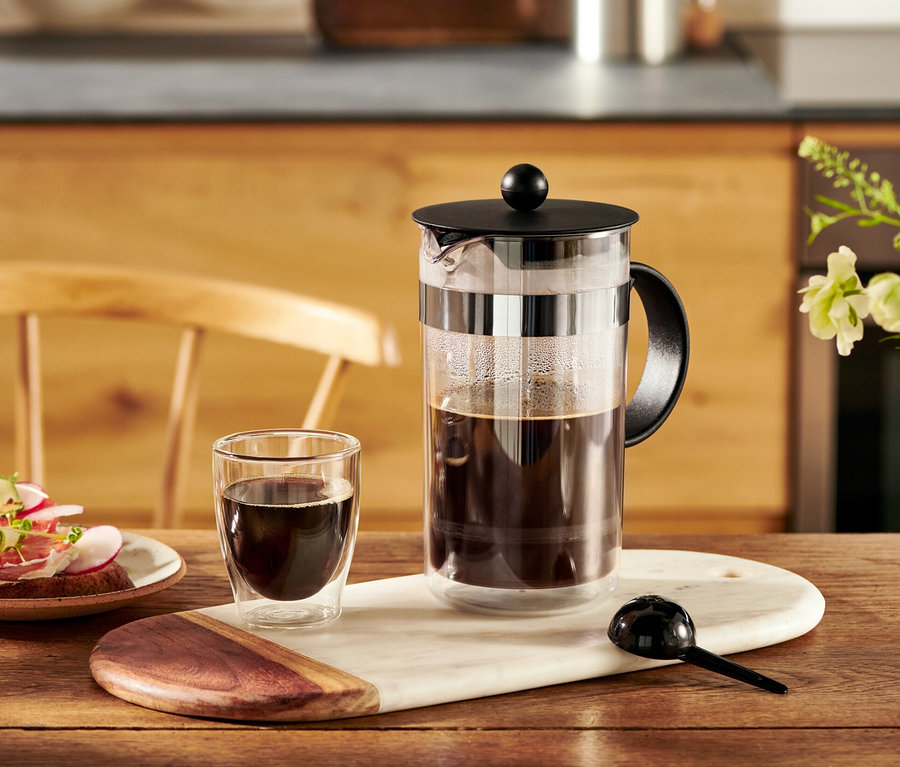 Eine Bodum® Siebstempelkanne, 1 l, steht auf einem Tisch neben einer Tasse Kaffee und einem Teller mit Brötchen.