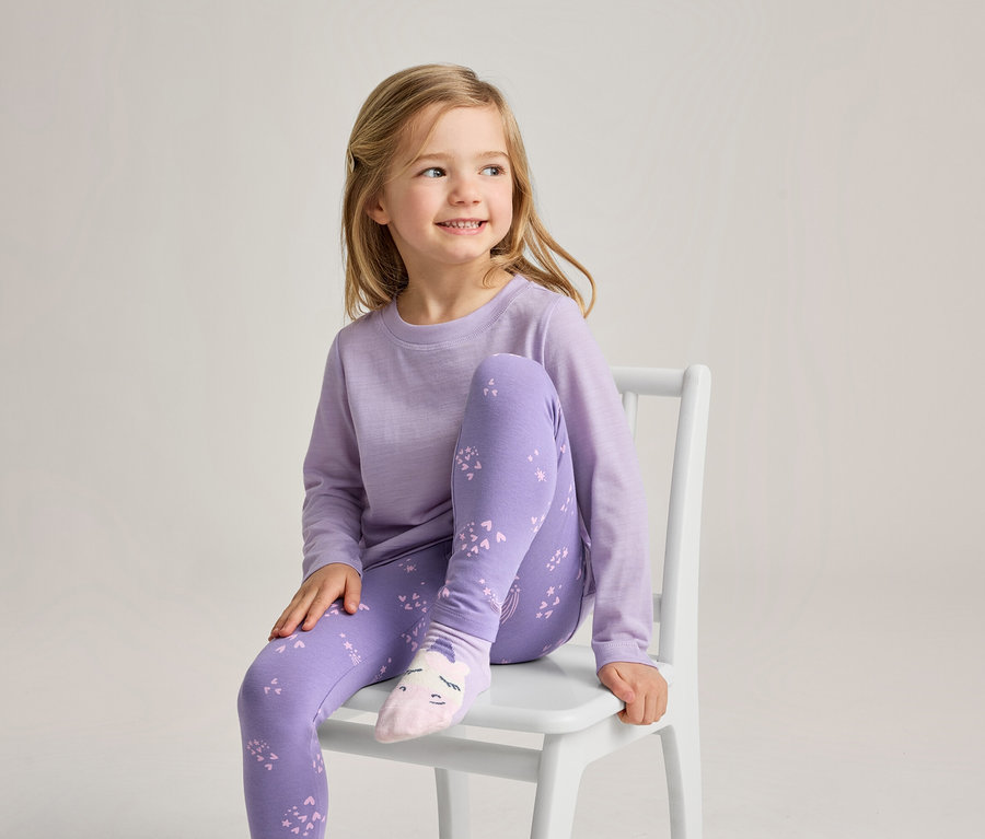 Ein junges Mädchen mit blonden Haaren sitzt auf einem weißen Stuhl, lächelt und blickt nach rechts. Sie trägt ein hellviolettes Langarmshirt, violette Leggings mit hellviolettem Muster und hellrosa Socken mit einem Gesichtsdesign.