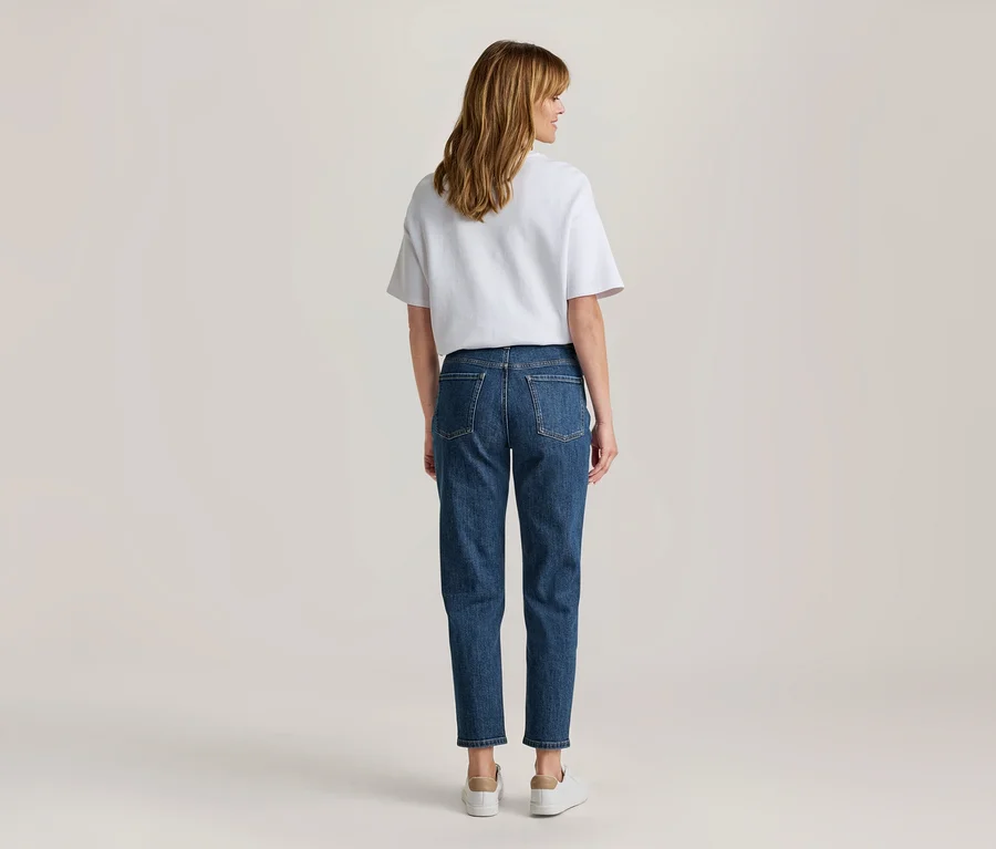 Frau von hinten, in weißem T-Shirt, blauen Jeans und weißen Sneakers, blickt über ihre rechte Schulter.