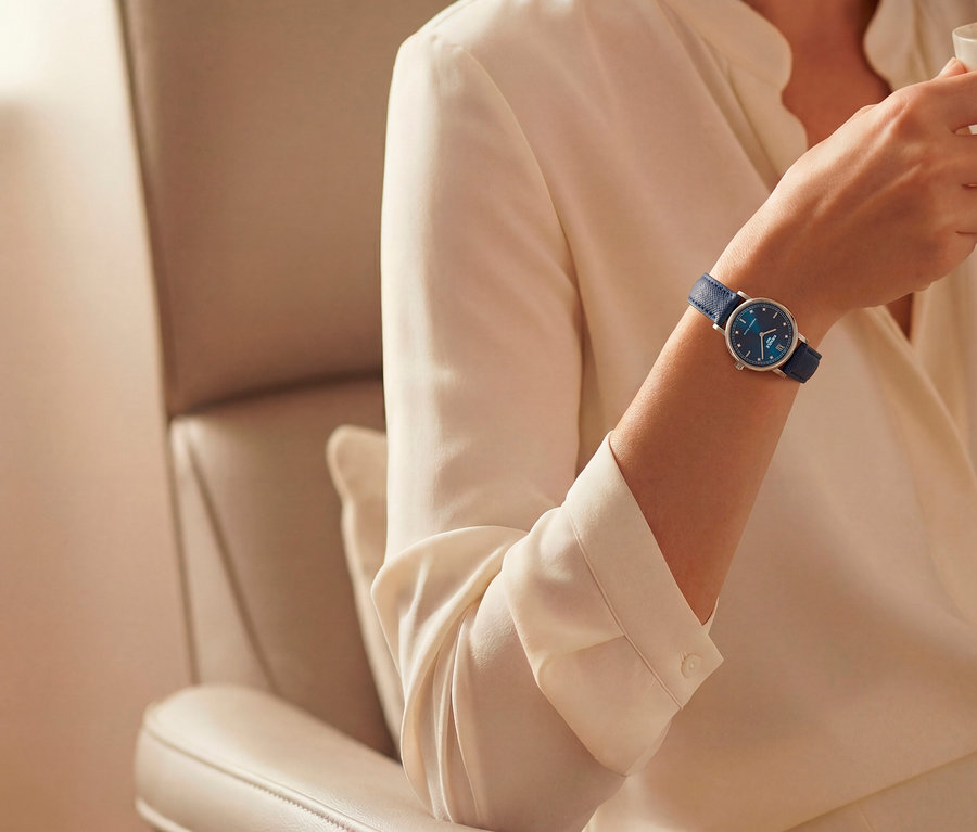 Arm einer Person mit cremefarbener Bluse, die eine Uhr mit blauem Zifferblatt und blauem Lederarmband trägt und eine Tasse hält.