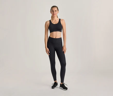 Eine Frau steht in einem schwarzen Sport-BH, schwarzen Leggings und schwarzen Sportschuhen vor einem hellen Hintergrund.