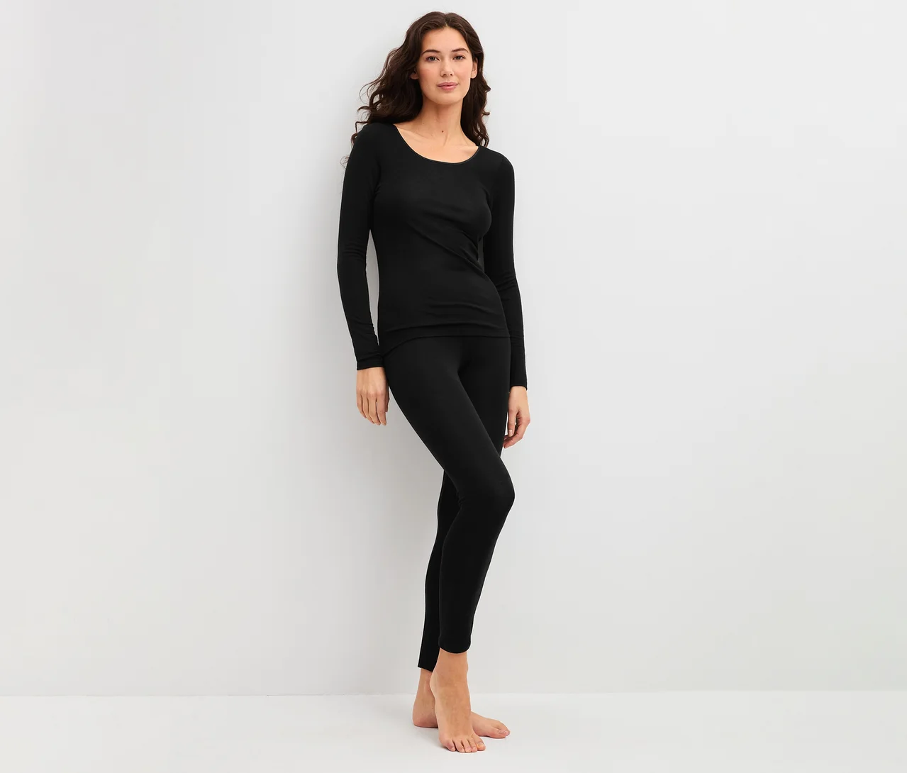 Frau mit langen dunklen Haaren trägt ein schwarzes CALIDA True Confidence Langarm-Shirt und schwarze Leggings, barfuß vor weißem Hintergrund stehend.