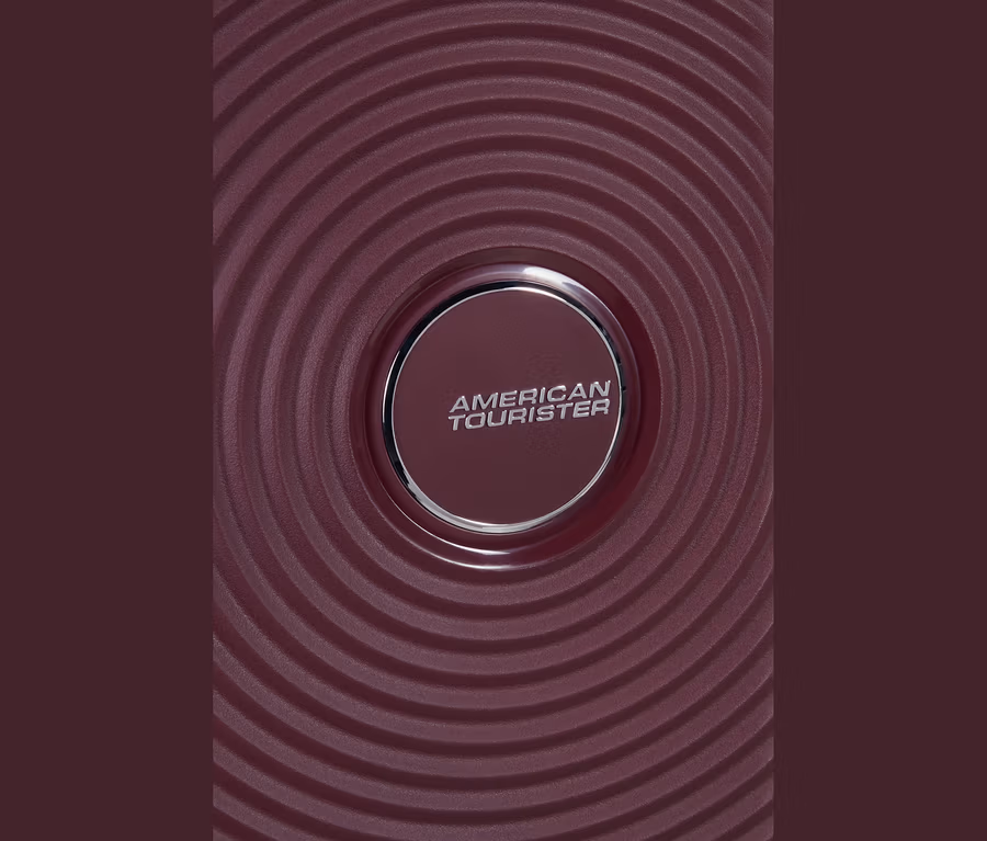 Nahaufnahme einer bordeauxroten Oberfläche mit konzentrischen kreisförmigen Rillen. In der Mitte befindet sich ein kreisförmiges, silberumrandetes Emblem mit dem Schriftzug „AMERICAN TOURISTER“.