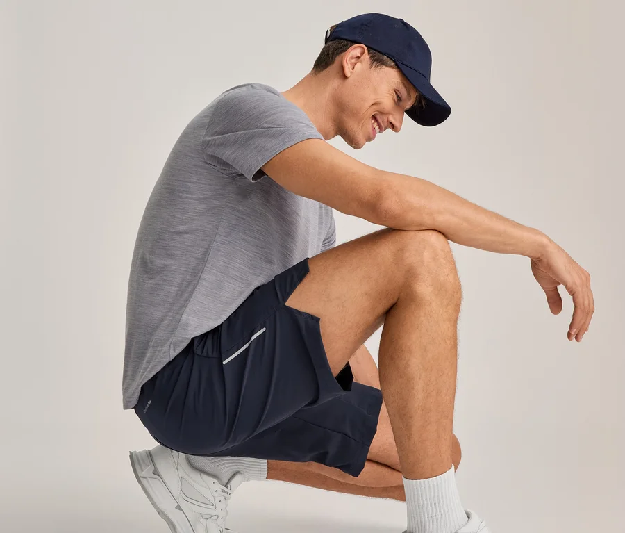 Mann hockt, lächelt und blickt nach unten. Er trägt ein graues T-Shirt, marineblaue Shorts mit einem weißen Streifen, eine marineblaue Baseballkappe, weiße Socken und weiße Turnschuhe.
