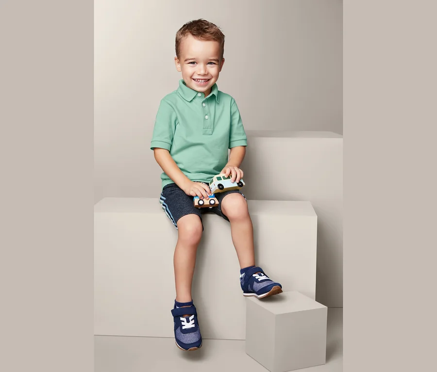 Ein Junge sitzt und hält zwei Spielzeugautos. Er trägt 2 Poloshirts und blaue Schuhe.