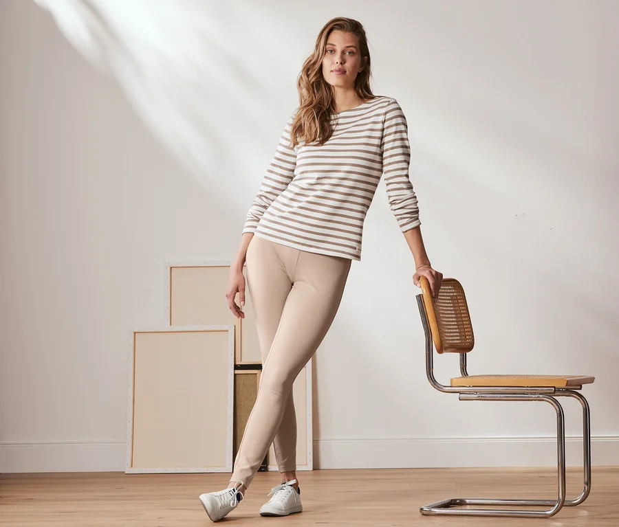 Frau posiert an einen Stuhl gelehnt in cremefarbene Basic-Leggings aus Heavy-Jersey und einem gestreiften Oberteil.