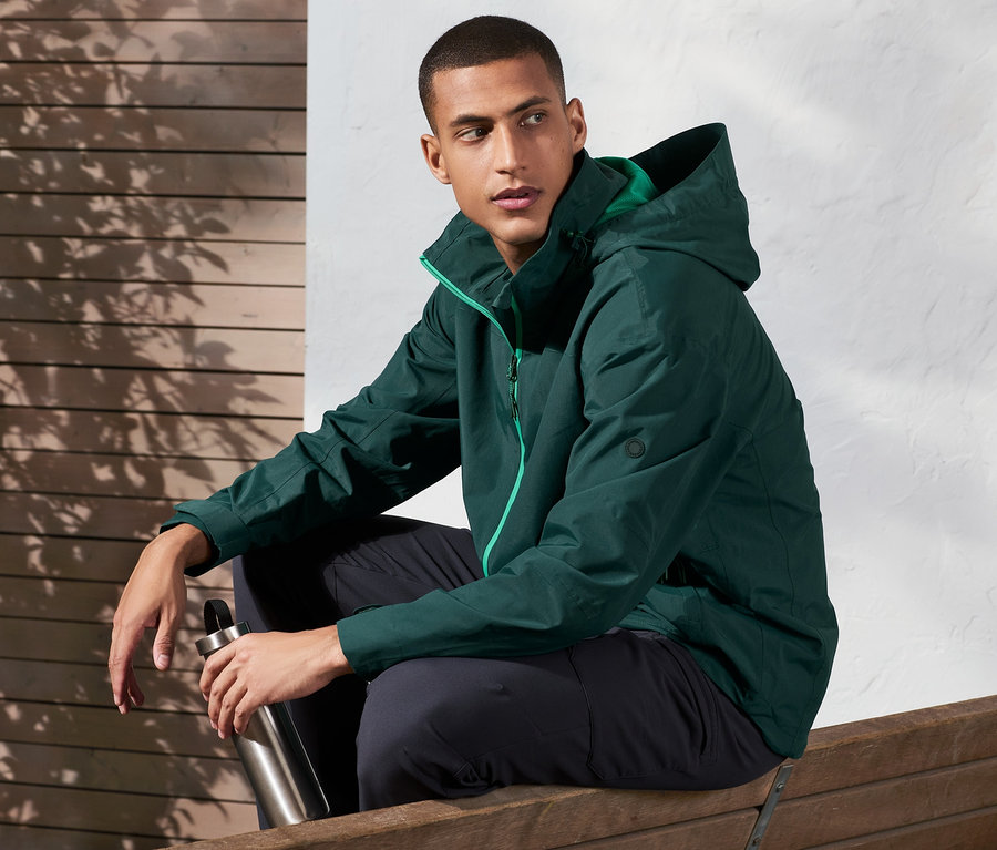 Mann sitzt auf einer Bank und hält eine Metallflasche. Er trägt eine Outdoorjacke.