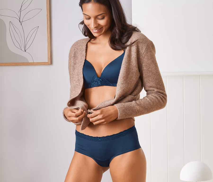 Frau öffnet braunen Cardigan, trägt blauen BH und 2 blaue Pantys.