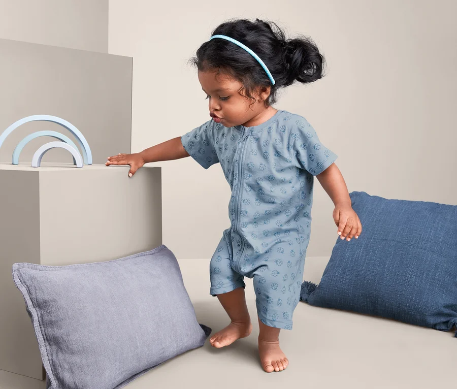 Kleines Mädchen in 2 Shorty-Pyjamas, grün-blau, steht neben blauem Spielzeug und Kissen.