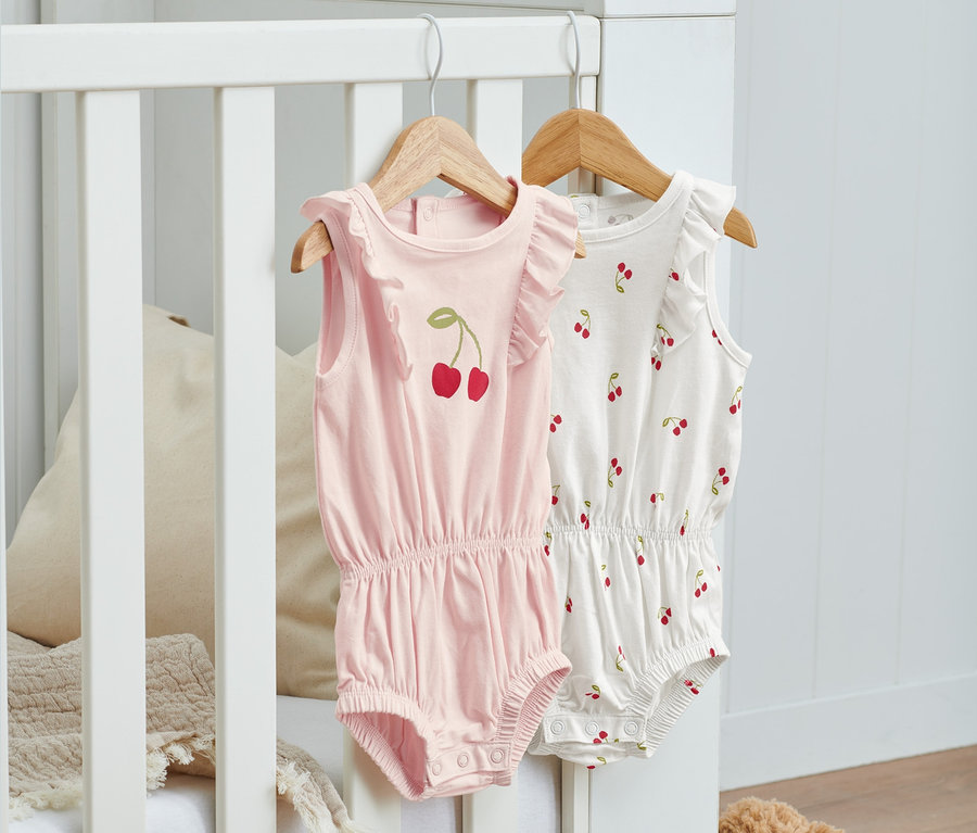 Zwei Bio-Baumwoll-Bodys mit Kirschmuster hängen auf Kleiderbügeln an einem Kinderbett.