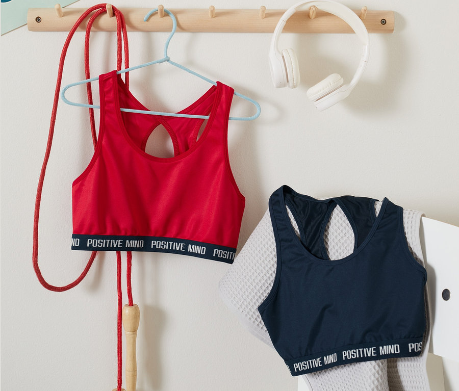 Ein rotes Sportbustier von 2 Sportbustiers hängt auf einem Bügel neben einem Springseil. Ein blaues Sportbustier von 2 Sportbustiers liegt auf einem grauen Stuhl neben einem Handtuch.