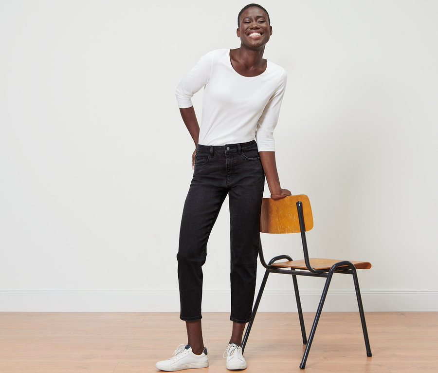 Eine lächelnde Schwarze lehnt an einem Stuhl und trägt Mom-Denim »Fit Nyla« und eine weiße Bluse.
