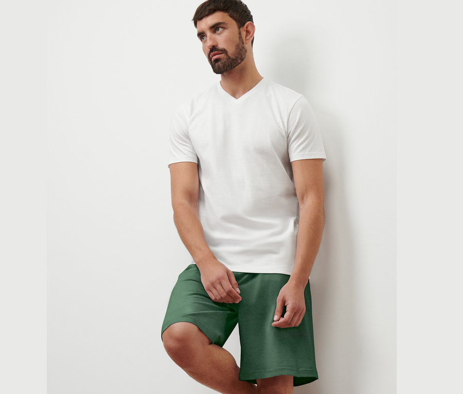 Mann lehnt an einer Wand, trägt ein weißes T-Shirt mit V-Ausschnitt und grüne Shorts. Er trägt 1 von 2 T-Shirts mit V-Ausschnitt.