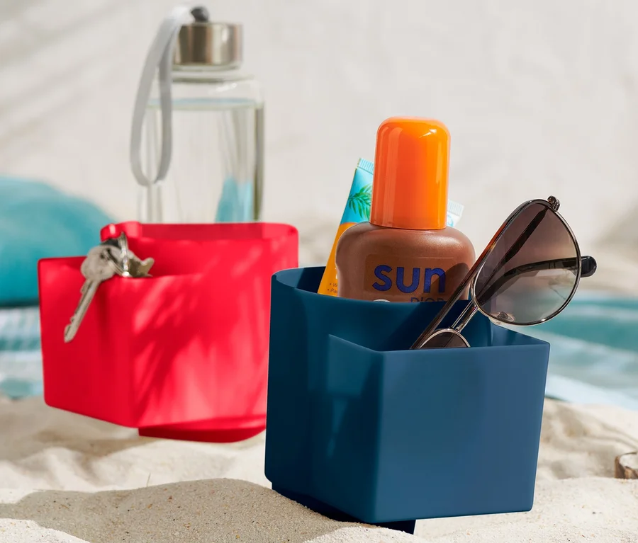 Zwei Strandbecher-Halter im Sand. Im blauen Halter befinden sich eine Sonnenbrille und Sonnencreme.