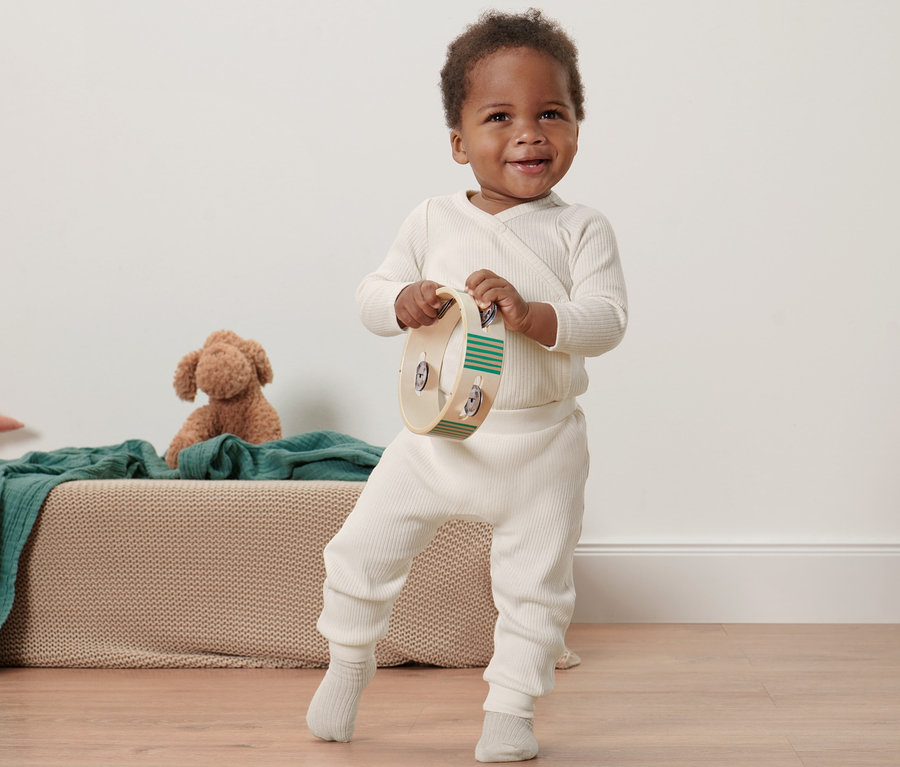Ein lächelndes, dunkelhäutiges Kleinkind hält ein Tamburin und steht in einer Baby-Strickhose.