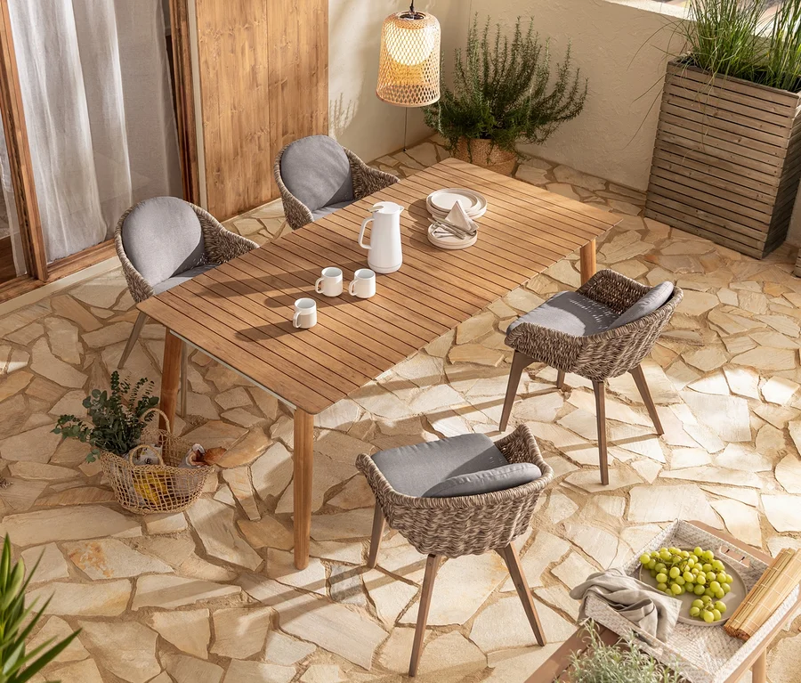 Terrasse mit Teak-Ausziehtisch, umgeben von Premium-Schalenstühlen mit Polyrattangeflecht und Gartenbank mit Rattan-Geflecht. Ein Pflanzkasten-Raumteiler »Tinus« strukturiert den Bereich. Akku-Outdoor-Hängeleuchten sorgen für Beleuchtung.
