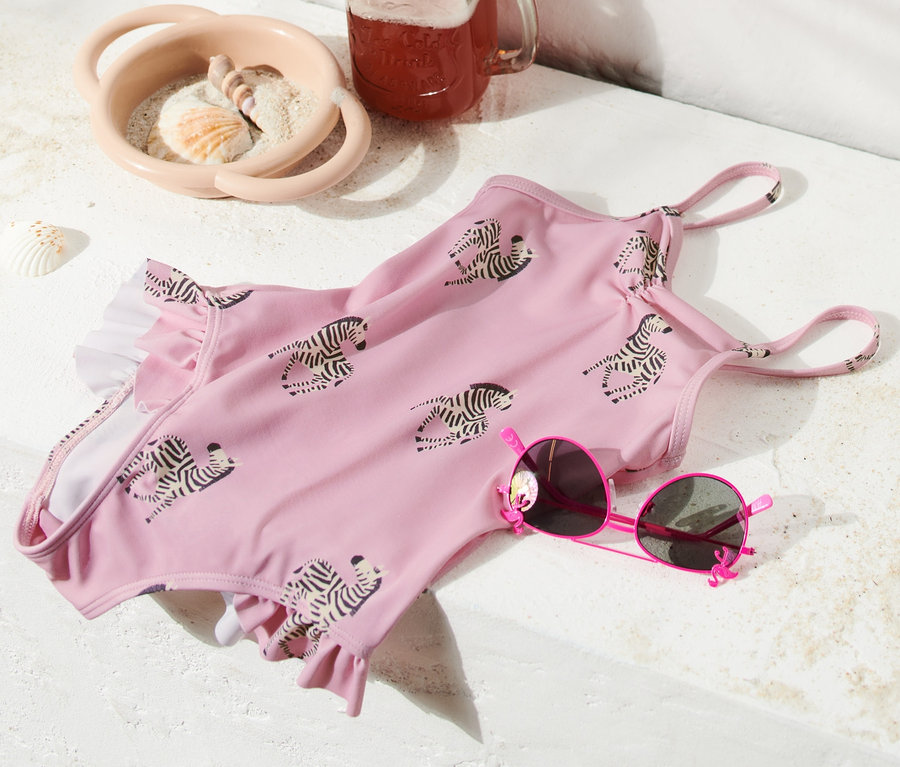 Ein rosafarbener Badeanzug mit Rüschen und Zebramuster liegt neben einer rosa Sonnenbrille, einem Getränk im Glas und einer Strandtasche.