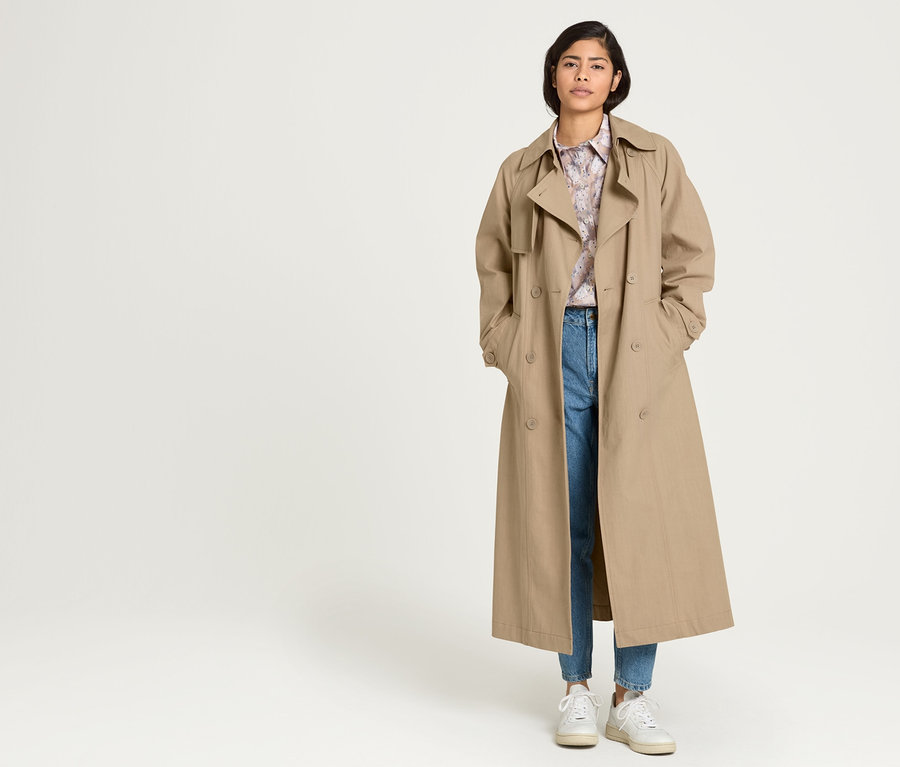 Frau steht mit Händen in den Taschen eines beigen NAH/STUDIO Trenchcoats aus recycelter Baumwolle, darunter eine bedruckte NAH/STUDIO Longbluse aus Bio-Baumwolle und eine hellblaue NAH/STUDIO Mom-Jeans aus recycelter Baumwolle.