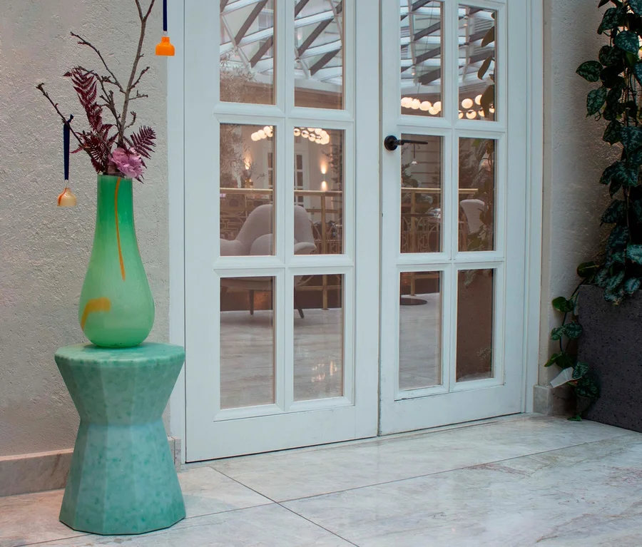 Blumen in Vase auf grünem Beistelltisch vor Glastür.