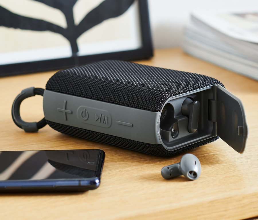 Schwarzer Bluetooth-Lautsprecher mit In-Ear-Kopfhörern auf einem Holztisch.