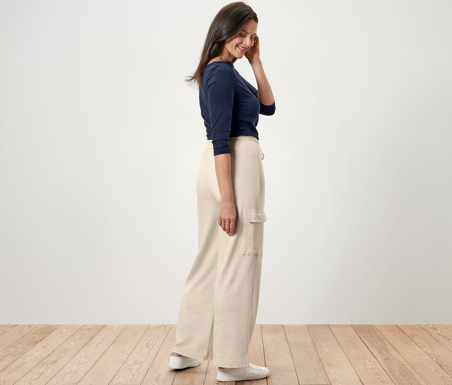 Eine Frau steht seitlich und trägt eine cremeweiße Sweathose mit Cargo-Taschen, ein blaues Shirt und weiße Turnschuhe.