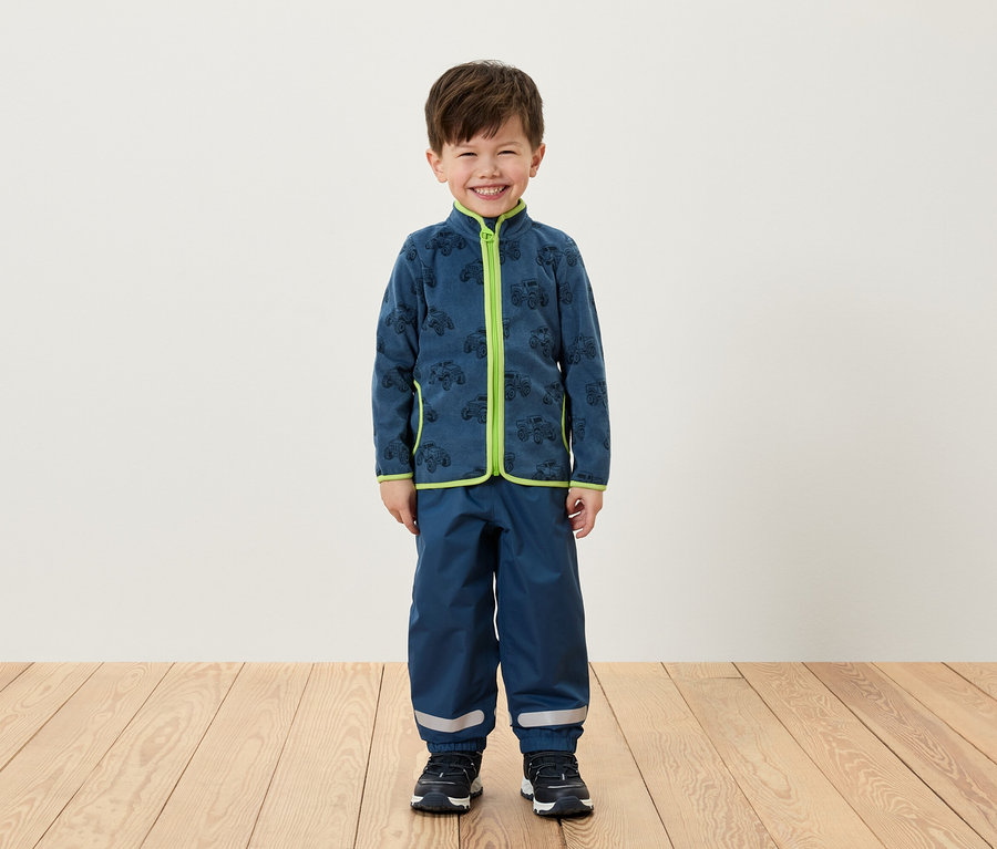 Ein Junge steht und trägt eine blaue Fleecejacke mit grünen Akzenten und eine blaue Hose.