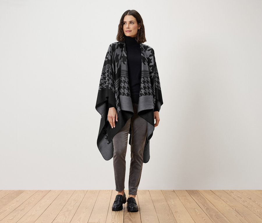 Eine Frau steht und trägt einen schwarzen Rollkragenpullover, graue Hosen und eine grau-schwarze Cape mit Hahnentrittmuster.