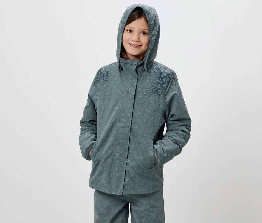 Ein lächelndes Mädchen posiert in einer Thermo-Regenjacke mit Kapuze und reflektierenden Elementen.
